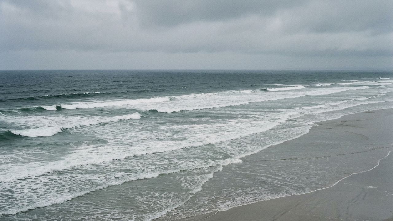 Successive lines of breaking surf running in toward a wide empty grey beach, low cloud
