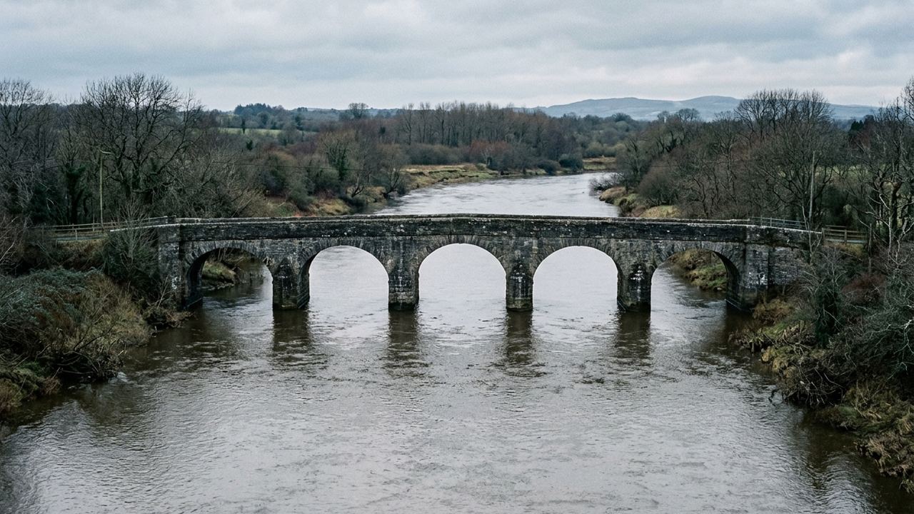 A stone bridge of several identical arches crossing a wide slow river, grey water, bare trees